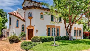 A two-story building with a white facade and red-tiled roof, featuring a central entrance with a wooden door, arched windows, and a small balcony. - Villa Blanca 4bd 3.5ba Private Pool and Parking (West Palm Beach)