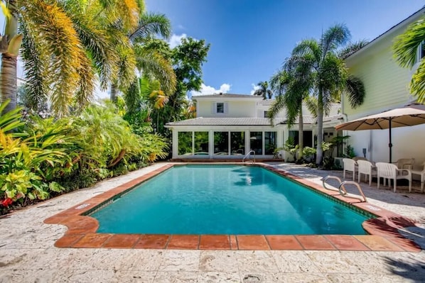 A swimming pool with a tiled deck, surrounded by palm trees and a covered patio area.