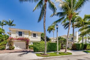 A two-story house with a white garage door, surrounded by palm trees and well-maintained hedges.