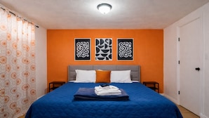 Quarto de hotel com uma parede laranja, uma cama com colcha azul, duas almofadas e um cobertor dobrado na cama.