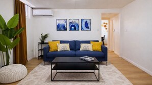 Uma sala de estar com um sofá azul, uma mesa de centro e uma planta em vaso. Há três quadros na parede.