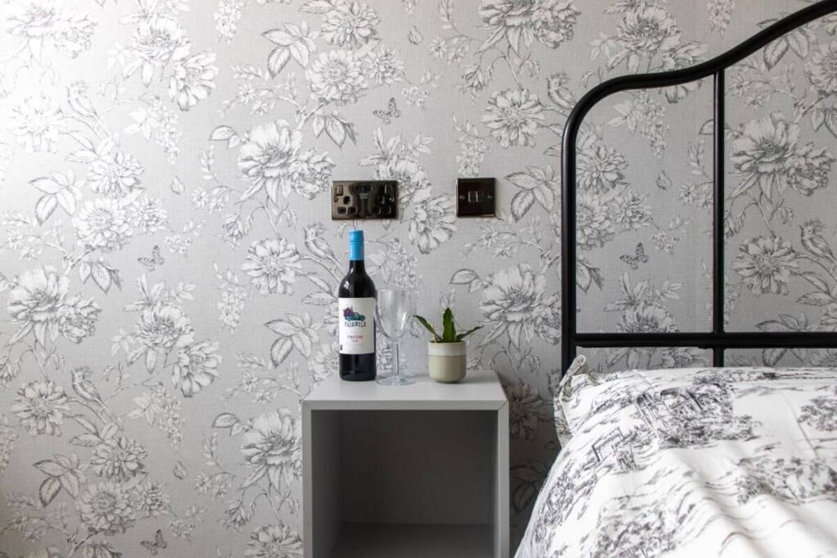 A bedroom with floral wallpaper, a bed with a patterned bedspread, a bedside table with a wine bottle and glass, and a small potted plant.