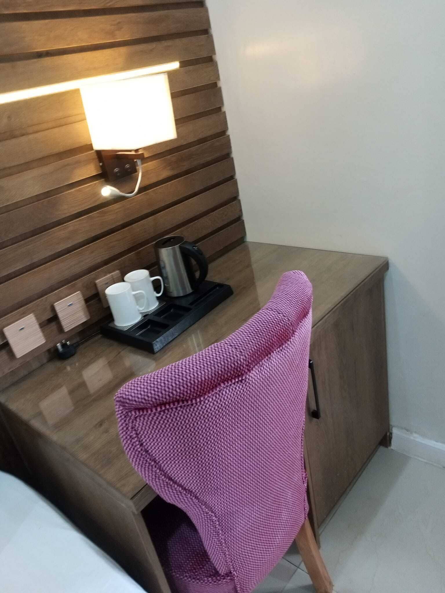 A small, modern room with a wooden desk, a pink chair, a coffee maker, and a wall-mounted light.