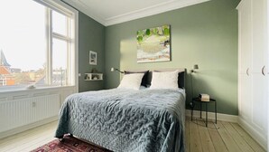 A bedroom with a large bed, a window with a view of buildings, a painting on the wall, and a small bedside table with a lamp.