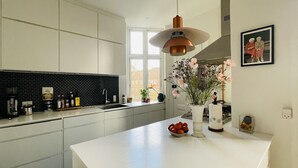 A modern kitchen with a white countertop, black backsplash, and a copper pendant light. There is a window with a view of buildings outside, and a framed picture on the wall.
