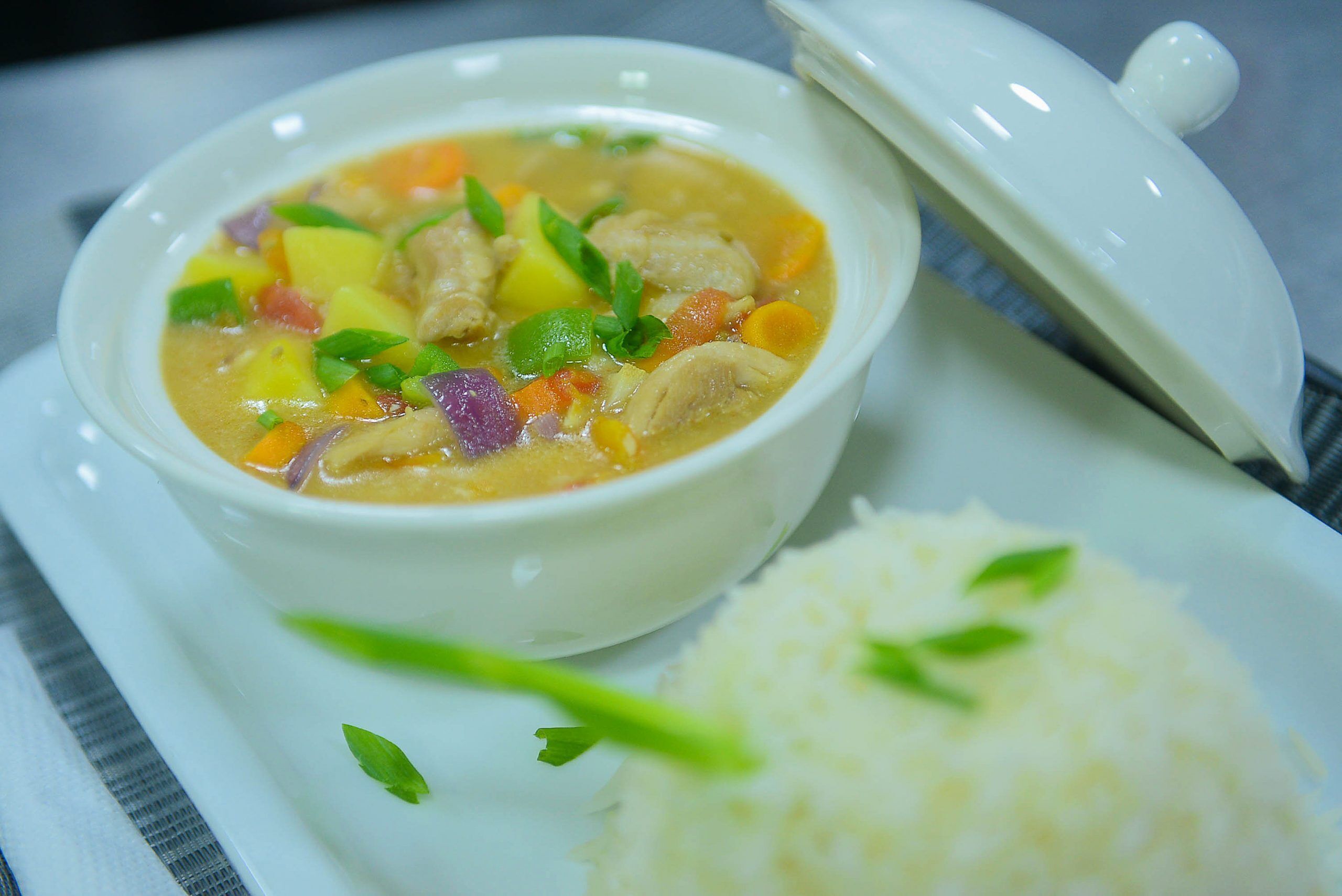 A bowl of soup with vegetables and rice on a white plate.