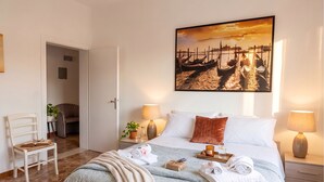 A neatly arranged hotel room with a bed, bedside lamps, a painting of boats, a chair, and a desk with a plant.