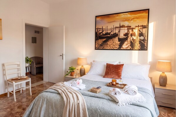 A neatly arranged hotel room with a bed, bedside lamps, a painting of boats, a chair, and a desk with a plant.