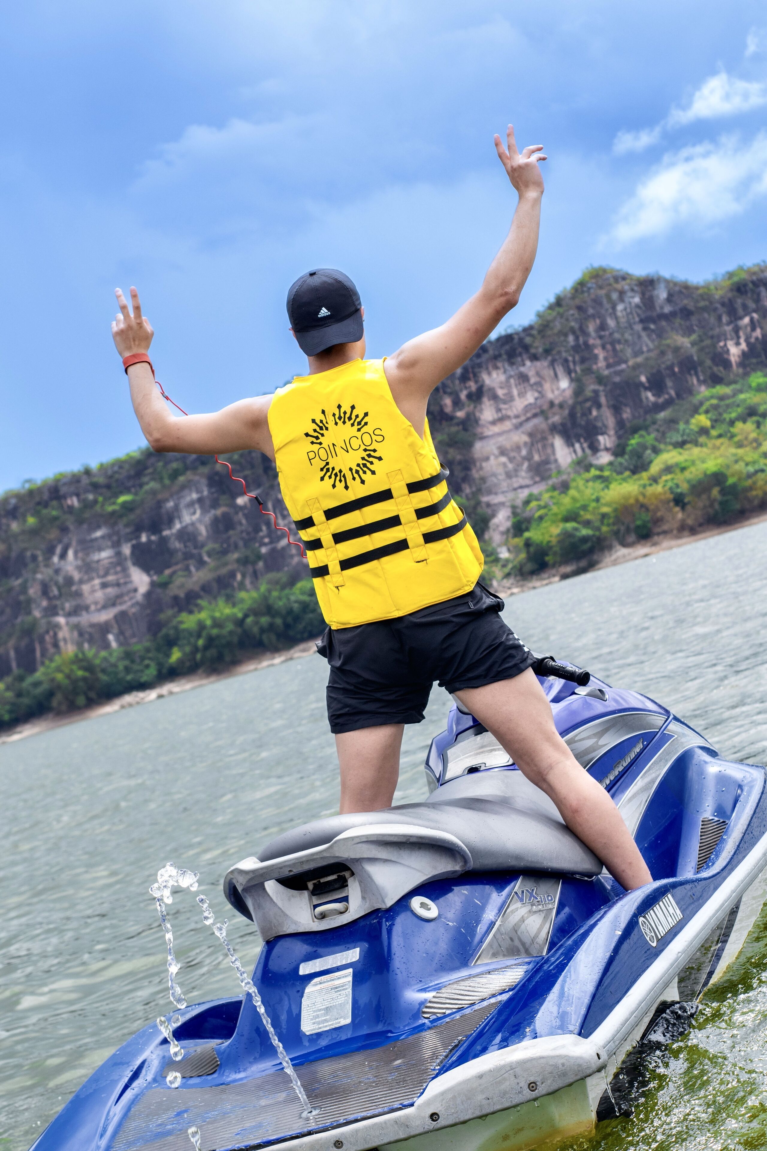 A person on a jet ski wearing a yellow life jacket with a sun design, standing on a blue and white jet ski with water splashing around.