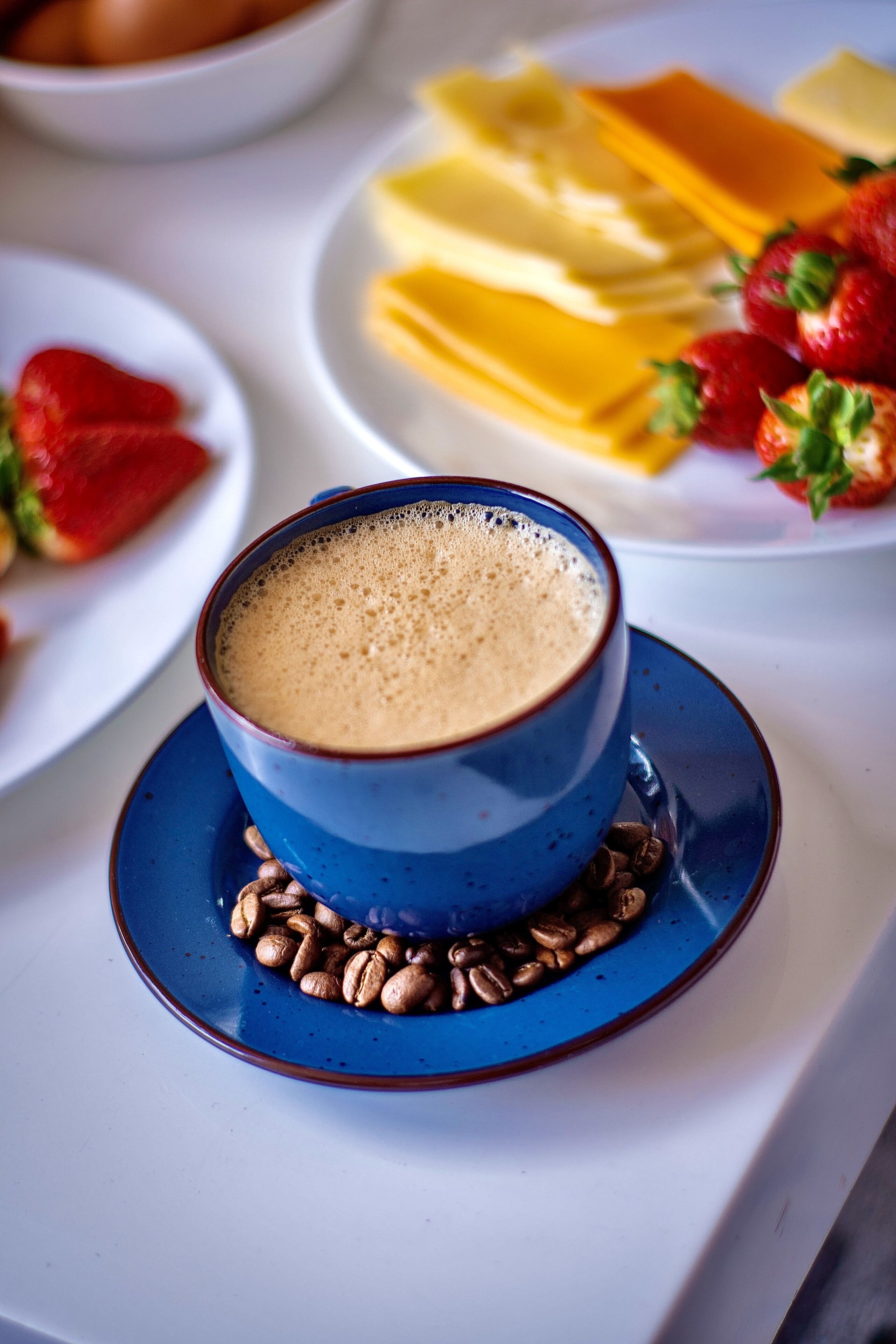 Una tazza di caffè su un piattino, con chicchi di caffè, un piatto con formaggio e fragole, e una ciotola con uova sullo sfondo.