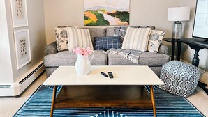 Ein Wohnzimmer mit grauem Sofa, gemusterten Kissen, einem weißen Couchtisch, einem blau-weißen Teppich, einem gerahmten Pflanzenbild und einem Gemälde an der Wand.