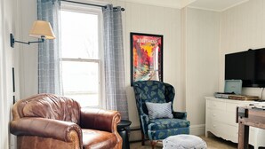 Ein Wohnzimmer mit einem braunen Ledersessel, einem blau gemusterten Sessel, einem kleinen Hocker, einem Fernseher auf einer Kommode und einem Poster an der Wand.