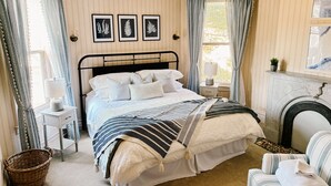 Ein ordentlich eingerichtetes Schlafzimmer mit einem Bett, Nachttischen, einem Sessel, einem Kamin und gerahmten Pflanzenbildern an der Wand.