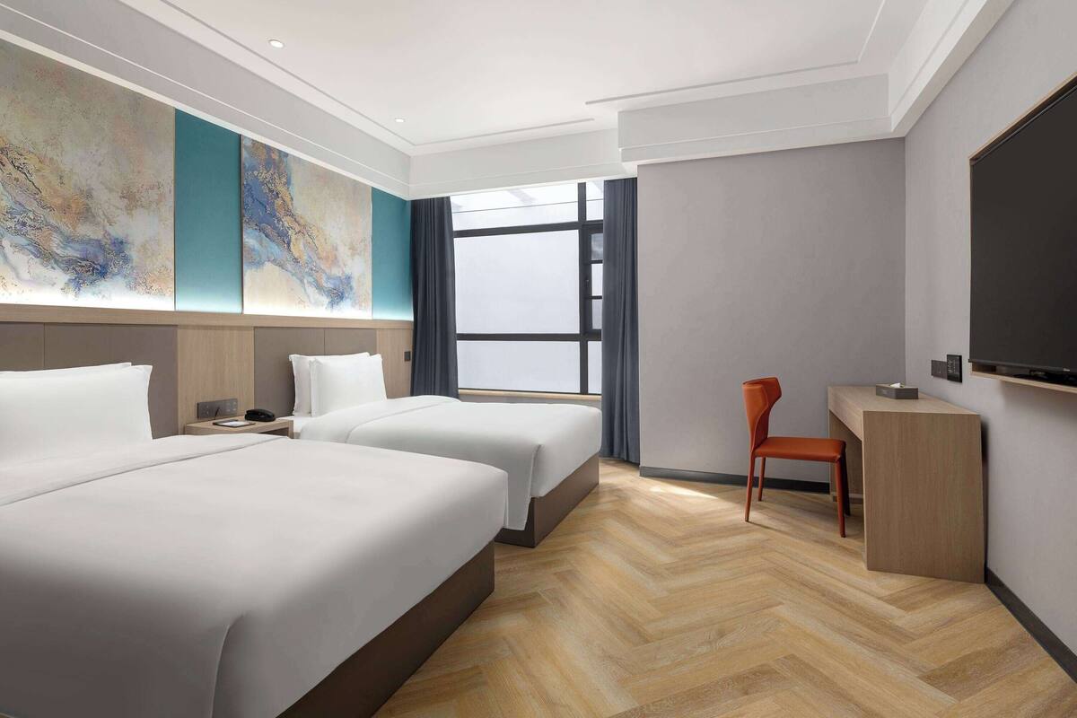 A hotel room with two beds, a desk, a chair, a TV, and a large abstract painting on the wall.