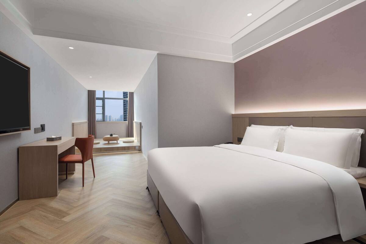 A modern hotel room with a large bed, a desk with a chair, and a view of the city through a large window.