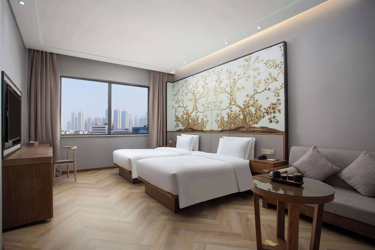 A hotel room with two beds, a sofa, a TV, and a large window with a city view.