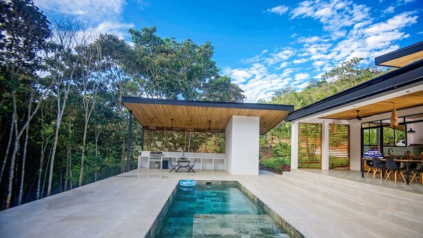 Un área de piscina moderna al aire libre con un jacuzzi integrado, rodeada por un porche cubierto con asientos y una zona de barbacoa.