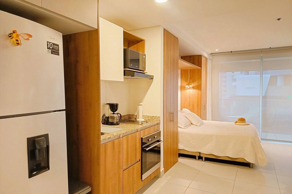 A compact hotel room with a kitchenette, a bed with a white cover, and a view of the outside through a large window.