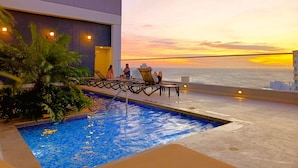 A rooftop pool with a view of the sunset over the ocean, surrounded by lounge chairs and a modern building. - Apartasuites Ambar Roca - Rodadero (Santa Marta)