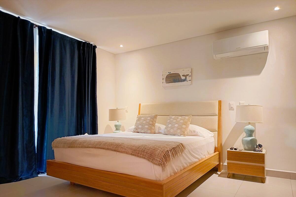 A hotel room with a wooden bed, two bedside tables with lamps, a wall-mounted air conditioner, and a window with dark curtains.