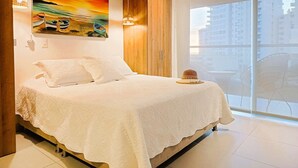 A bedroom with a bed, a painting of a sunset over water, and a view of buildings through a large window. - Apartasuites Ambar Roca - Rodadero (Santa Marta)
