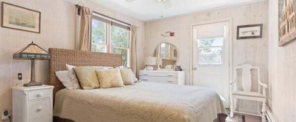 Un dormitorio con cama, mesita de noche, cómoda, espejo, silla y ventana con cortinas.