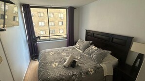 Een slaapkamer met een bed, een nachtkastje, een lamp, een raam met gordijnen en uitzicht op gebouwen.