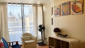 Een woonkamer met een bank, fauteuil en een bijzettafeltje met decoratieve objecten. Er staat een ventilator en een raam met gordijnen.
