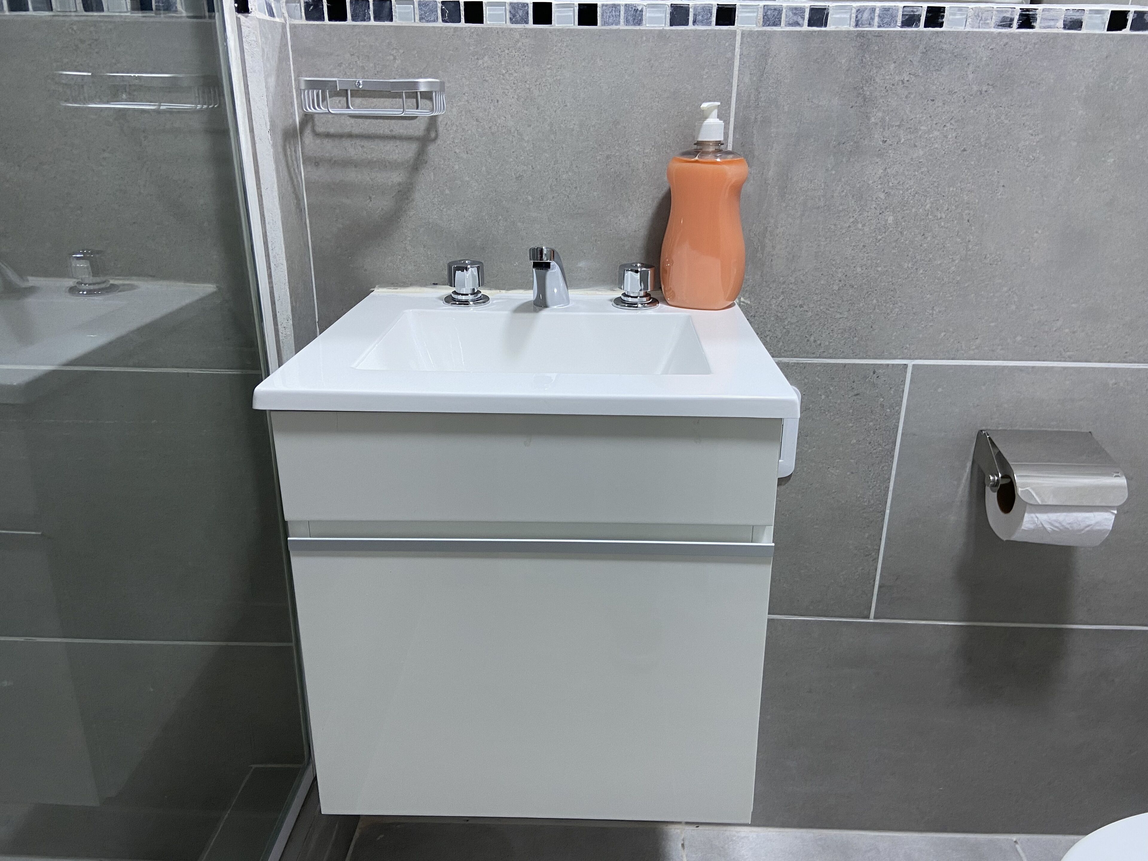 Un baño con un lavabo blanco, dos grifos, dispensador de jabón y soporte para papel higiénico.