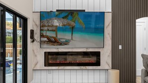 Uma sala de estar moderna com uma grande televisão de tela plana fixada em uma parede branca, uma lareira abaixo e uma janela ampla com vista para o exterior.