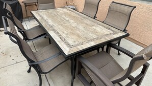 Una mesa rectangular al aire libre con tapa de madera desgastada y estructura de metal negro, rodeada por seis sillas con asientos y respaldos de fibras trenzadas.
