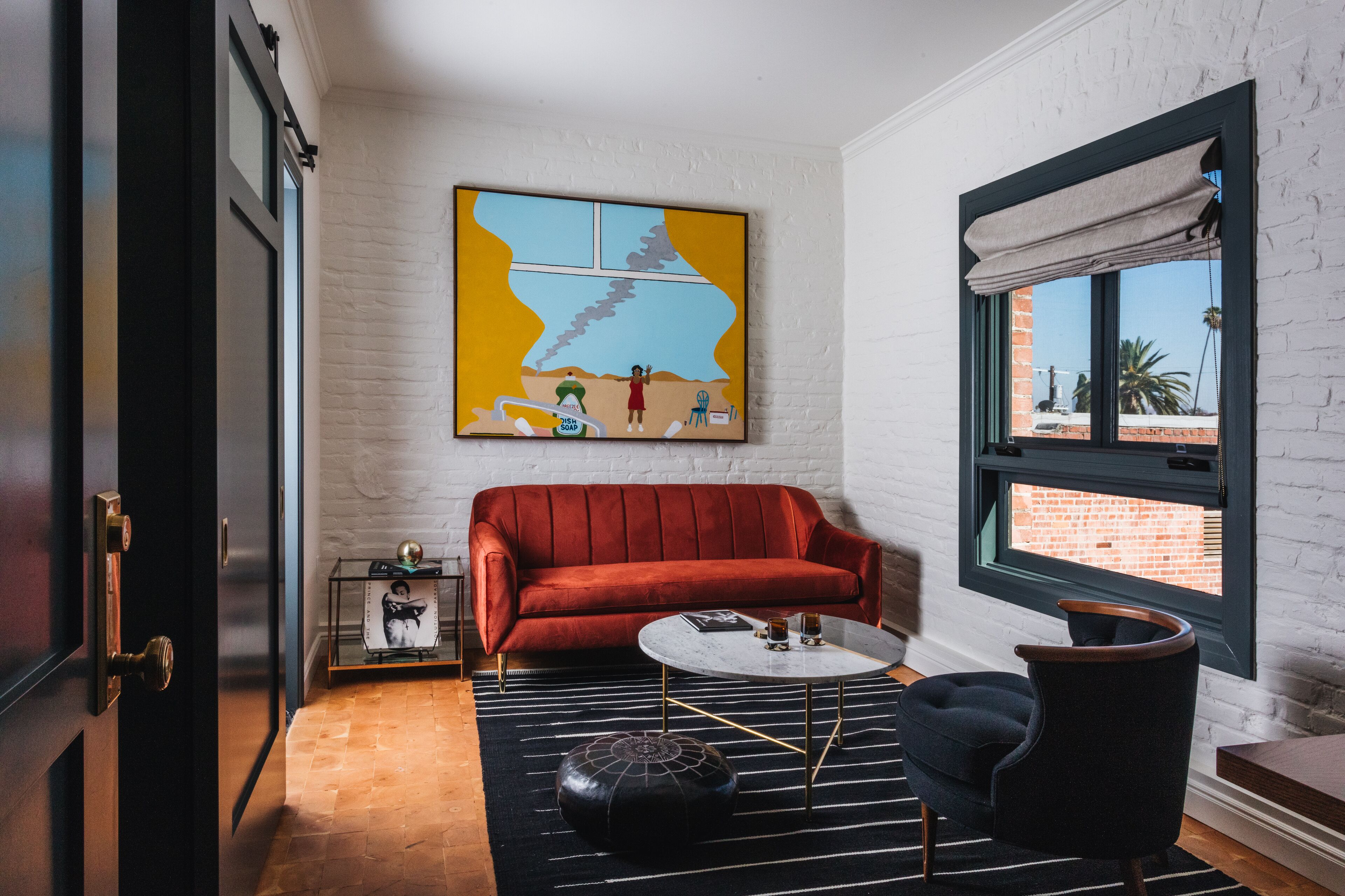 Ein modernes Wohnzimmer mit einem roten Sofa, einem runden Couchtisch und einem schwarzen Hocker. An der Wand hängt ein farbenfrohes abstraktes Gemälde, und es gibt ein Fenster mit Blick auf ein Backsteingebäude.
