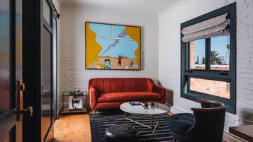 Ein modernes Wohnzimmer mit einem roten Sofa, einem runden Couchtisch und einem schwarzen Hocker. An der Wand hängt ein farbenfrohes abstraktes Gemälde, und es gibt ein Fenster mit Blick auf ein Backsteingebäude.