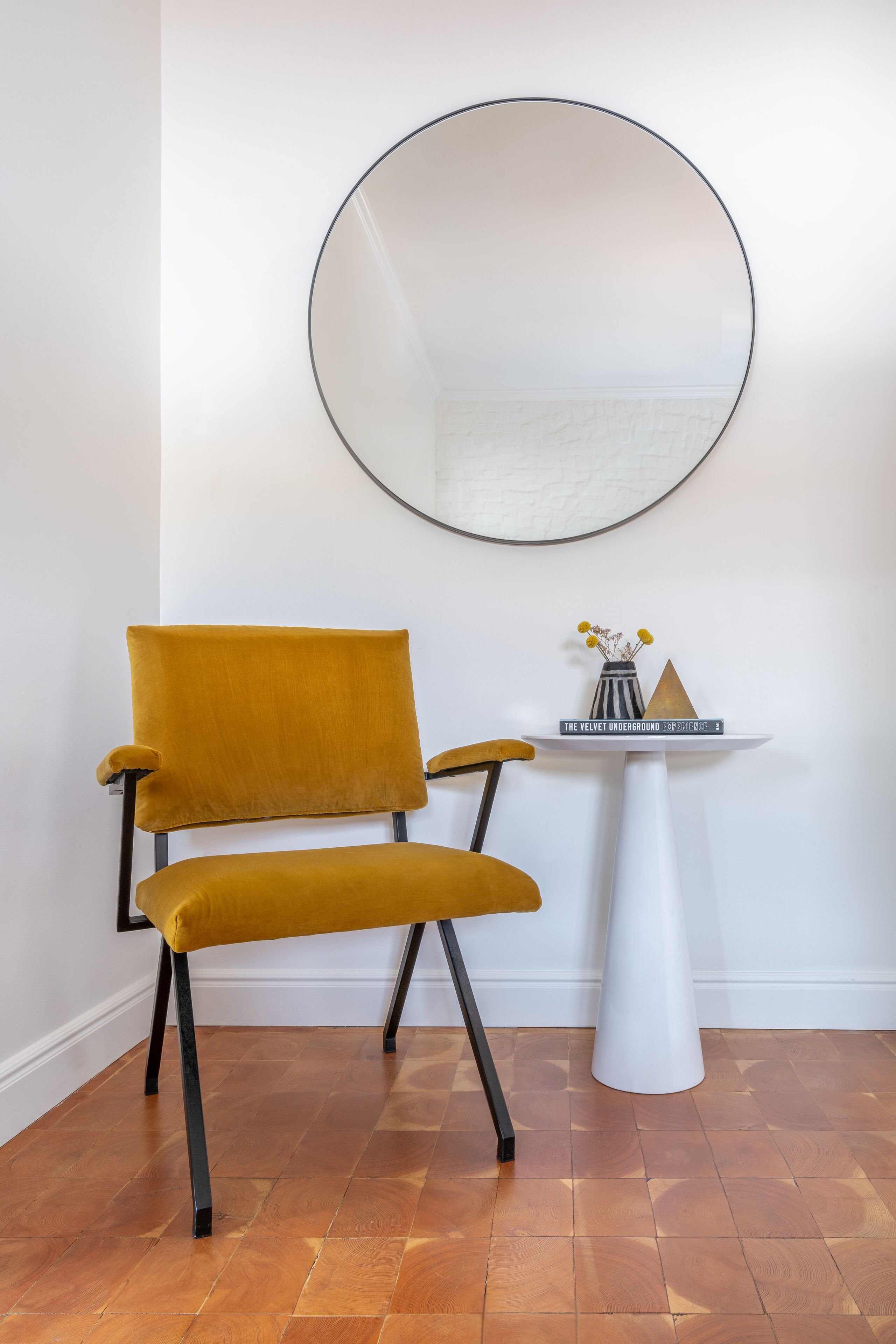 A mustard-colored chair with black legs, a white pedestal table with a mirror, and a small shelf with decorative items.