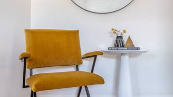A mustard-colored chair with black legs, a white pedestal table with a mirror, and a small shelf with decorative items.