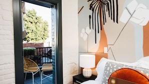 A hotel room with a bed, a nightstand with a lamp, a chair, and a view of the outside. - Gold-Diggers Hotel (Los Angeles)