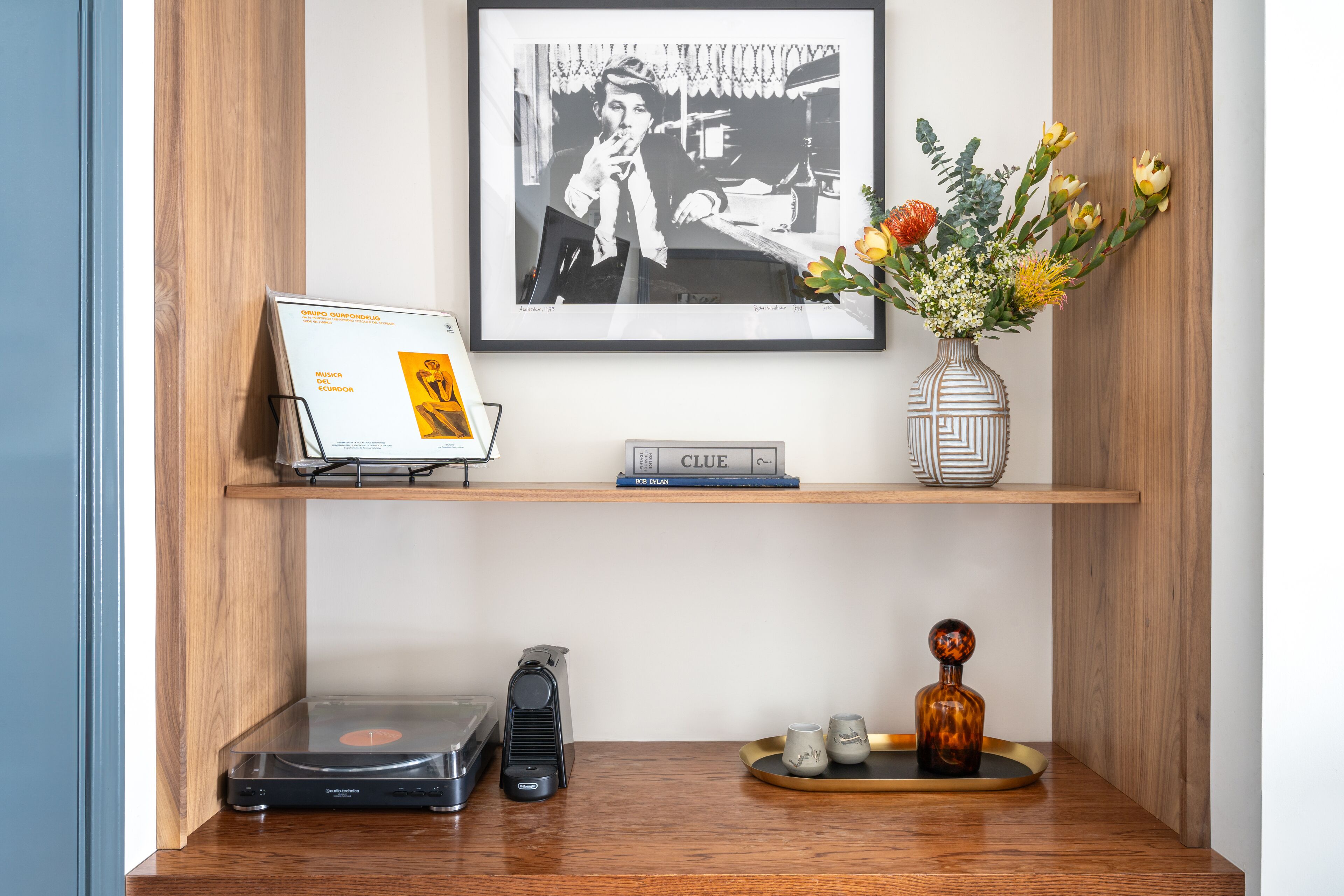 A wooden shelf with a framed black and white photograph, a small vase with flowers, a book titled 'CLUE', and a turntable.