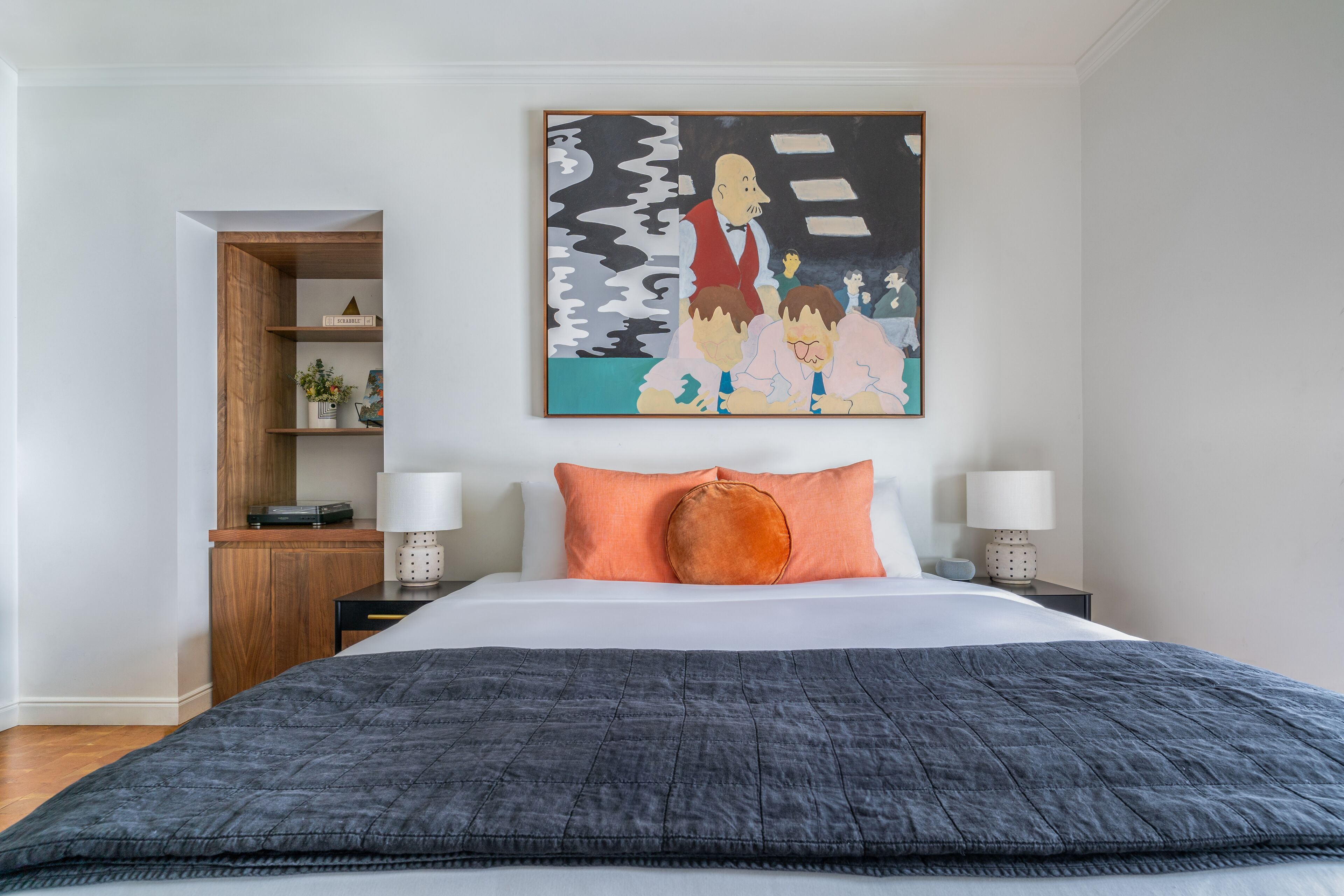 Un dormitorio con una cama, dos mesitas de noche, una mesita auxiliar, un estante de madera y un cuadro abstracto de gran tamaño.