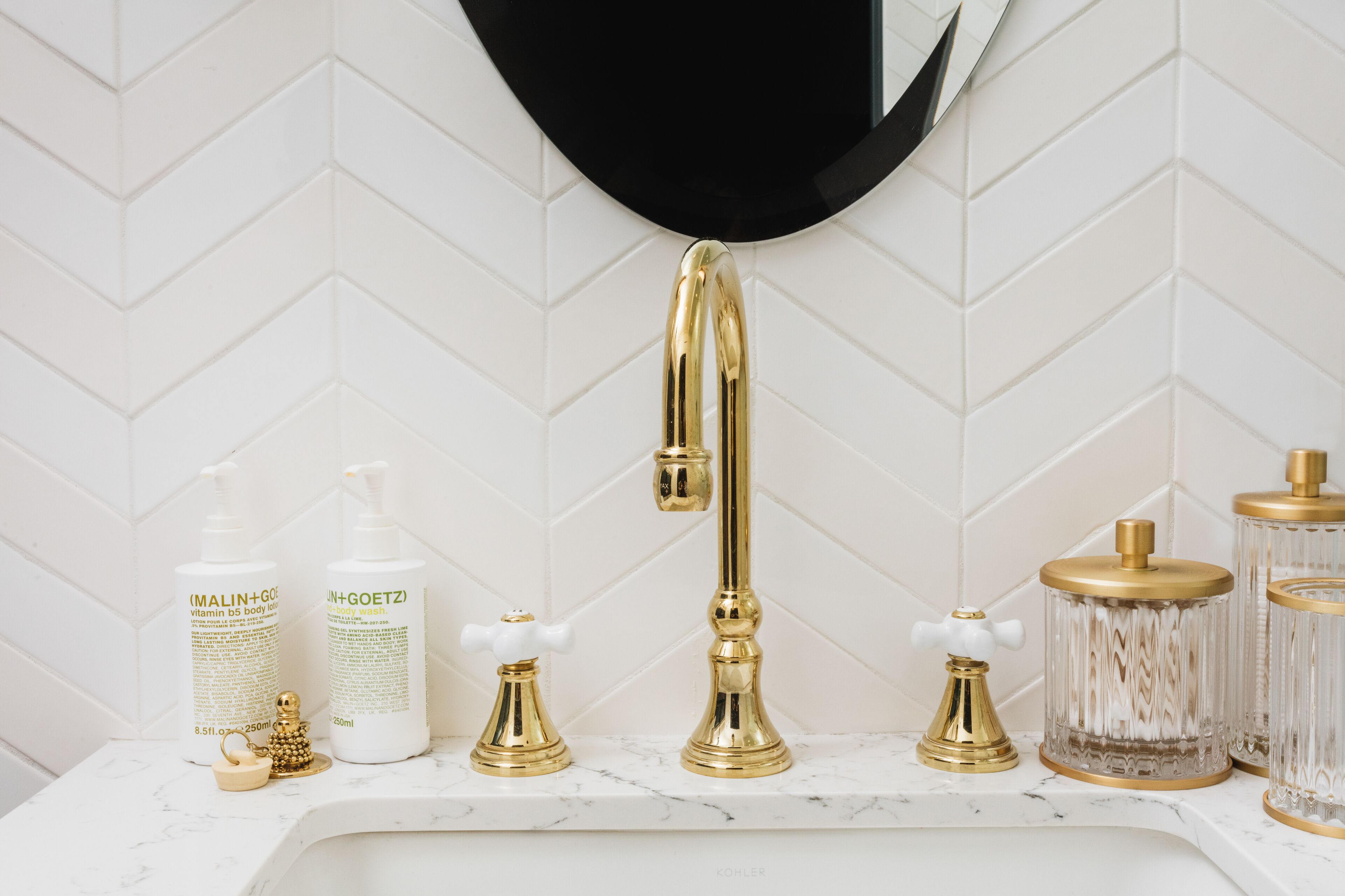 A bathroom vanity with a gold faucet, two bottles of body wash, a soap dispenser, and a glass container with a gold lid.