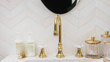 A bathroom vanity with a gold faucet, two bottles of body wash, a soap dispenser, and a glass container with a gold lid.