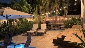 Un patio al aire libre con piscina, sillas de descanso, una mesa de madera y luces colgantes.