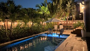 Una piscina con sillas y una sombrilla, rodeada de exuberante follaje e iluminación con luces colgantes.
