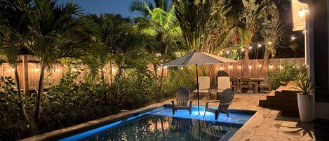 A swimming pool with chairs and an umbrella, surrounded by lush greenery and string lights.