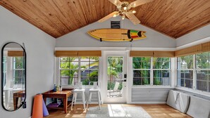 Una habitación con techo de madera, una tabla de surf, un espejo, equipo de ejercicio y ventanas grandes.