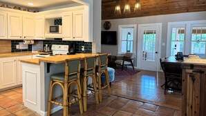 Una cocina con armarios blancos, una isla de madera con taburetes de bar, una zona de comedor con mesa y sillas, y un techo de madera con luces colgantes.