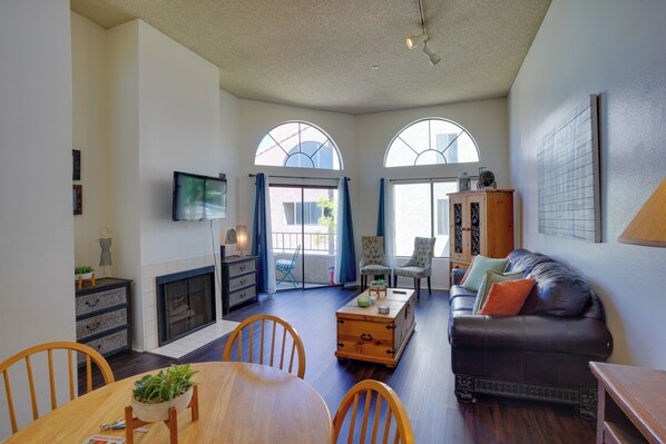 A spacious living room with a fireplace, a dining area with wooden furniture, and a large window with arched top. - 1 Mi to the Ocean: Long Beach Condo w/ Pool Access (Long Beach)