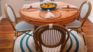 A dining area with a round wooden table, four chairs, a bowl of fruit, and a window with blinds. - Charming Cottage Downtown Hattiesburg (Hattiesburg)