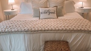 A neatly made bed with a patterned bedspread, two bedside lamps, and a pillow with the text "BE OUR GUEST". - Charming Cottage Downtown Hattiesburg (Hattiesburg)