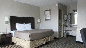 Une chambre d’hôtel comprenant un lit, des lampes de chevet, un tableau encadré, une salle de bain avec un lavabo et un miroir, et une poubelle.