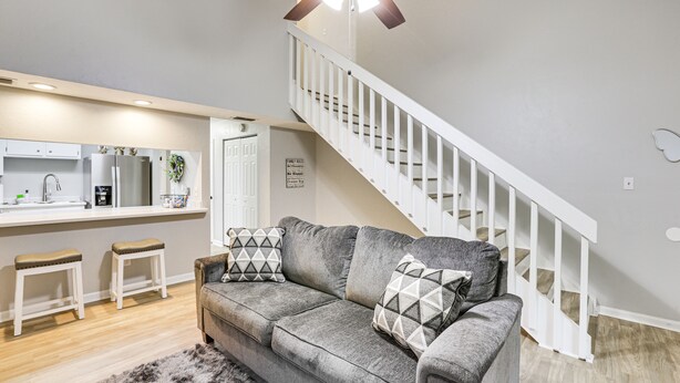 A modern living room with a gray sofa, two white bar stools, a kitchen area with a white countertop, and a white staircase. - Updated Tallahassee Townhome: 3 Mi to Downtown! (Tallahassee)